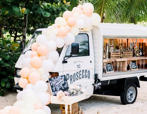The image features a stylish, white Prosecco van decorated with a large array of white and peach-colored balloons