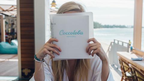 This image shows a woman sitting at a bar and grill named Coccoloba, holding a menu which covers her face