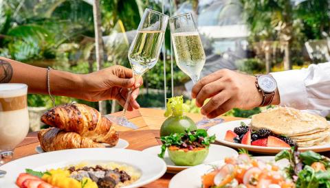 The image shows a luxurious outdoor dining setting featuring two people toasting with champagne glasses