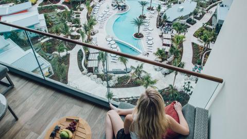 The image shows a person sitting on a balcony, overlooking a tropical resort pool area