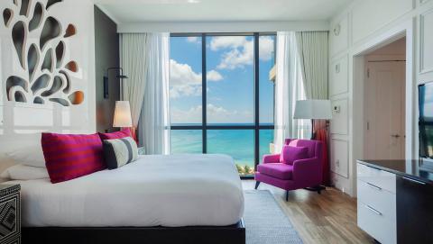 This image shows a stylish modern hotel room with a large window offering a beautiful view of the ocean and sky
