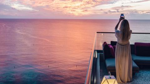The image depicts a woman standing on a balcony, capturing a photo of a stunning seaside sunset with her smartphone
