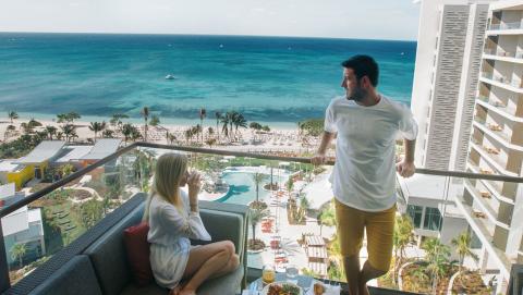 The image shows a man and a woman on a balcony overlooking a beautiful beach scene