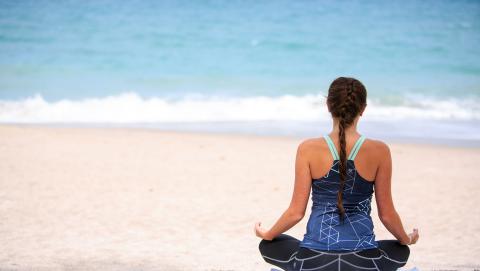 The image shows a woman seated in a meditative pose on a sandy beach, facing the ocean