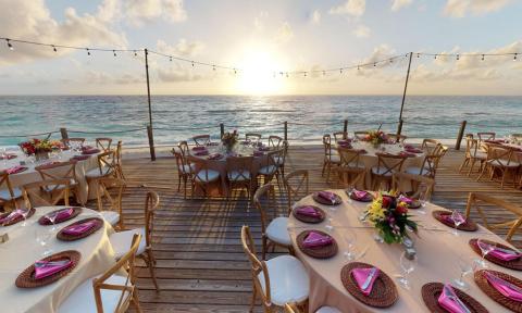 The image shows an outdoor dining setup on a wooden deck overlooking the sea during sunset
