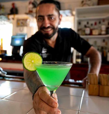 This image showcases a smiling bartender holding out a bright green cocktail garnished with a slice of lime