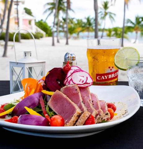 The image features a colorful plated meal set outdoors, likely at a beachfront location, given the visible sand and palm trees in the background