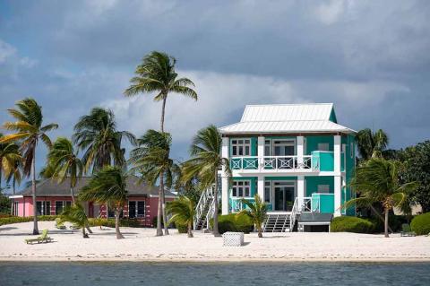 This image shows a beautiful coastal scene featuring two buildings on a white sandy beach, adorned with palm trees swaying in the wind