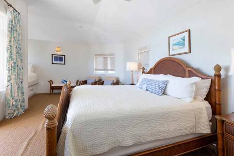 The image shows a well-lit and airy bedroom featuring a large bed with a white quilt and multiple pillows