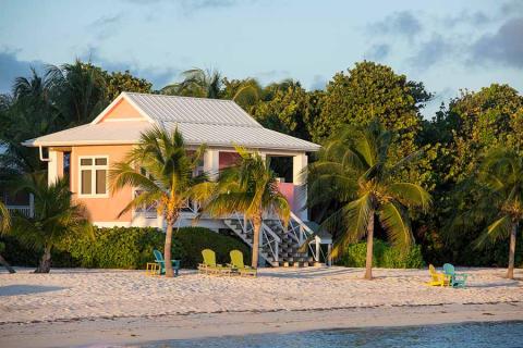 This image depicts a serene beachfront scene with a charming pink house surrounded by lush greenery and palm trees