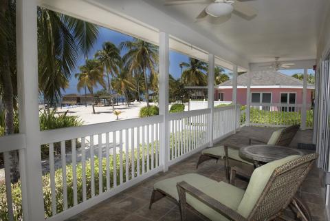 This image shows a tranquil veranda with a seating area that includes wicker chairs and a table, looking out over a beach scene