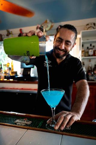 This image features a bartender smiling as he pours a bright blue cocktail from a green cocktail shaker into a martini glass at a bar