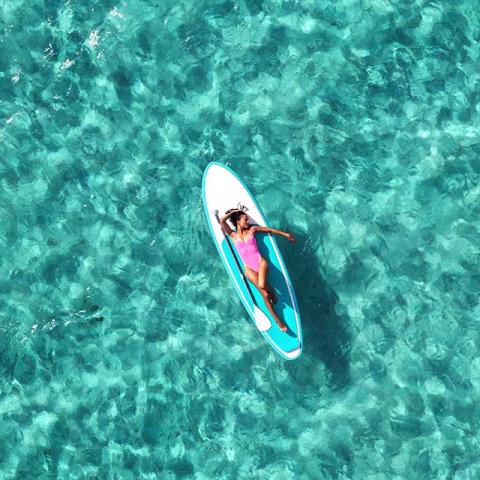 This image shows an aerial view of a person lying on a paddleboard in clear, turquoise water