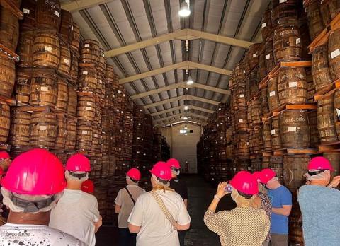 This image shows a group of people, likely on a guided tour, inside a large storage facility filled with stacked wooden barrels