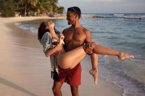 This image shows a joyful scene on a beach where a man is carrying a woman in his arms