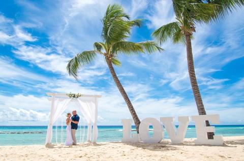 The image depicts a beautiful beach wedding scene under a clear blue sky