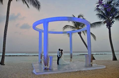 This image features a romantic beach setting during sunset where a couple is standing under a decorative, oval-shaped arch