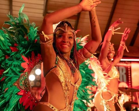 The image features two women dressed in vibrant and elaborate carnival costumes, performing or dancing at an event