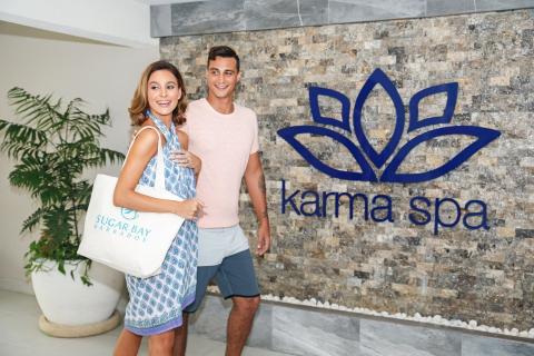 This image shows a man and a woman standing together in front of a stone wall with the logo of "Karma Spa" on it
