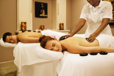 The image shows a spa scene where a man and a woman are receiving hot stone massages