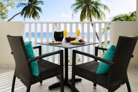 This image shows a tranquil outdoor breakfast or dining setting on a balcony overlooking a beautiful beach scene