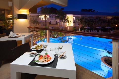 This image features a dining setup on an outdoor terrace with a picturesque view of a brightly lit pool at dusk