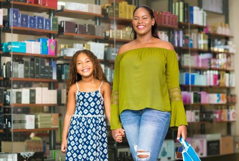 This image shows two individuals, likely a mother and daughter, in a retail store with shelves stocked with various boxed products in the background