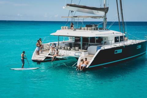 The image shows a group of people engaged in various leisure activities on and around a large catamaran anchored in a calm, turquoise sea