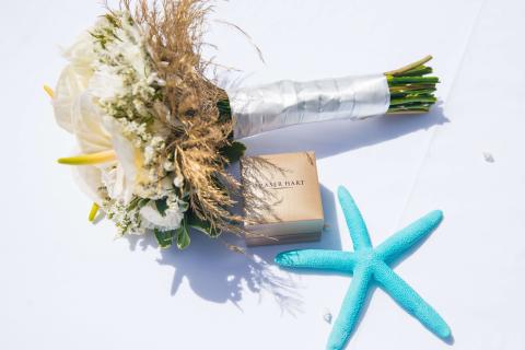 The image features a beautiful bridal bouquet with white flowers and greenery, lightly accented with some dried grasses for a rustic or beachy addition