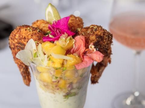This is an image of a gourmet dish featuring what appears to be coconut-crusted shrimp elegantly presented atop a glass filled with a creamy, layered sauce or dip, possibly containing herbs and spices