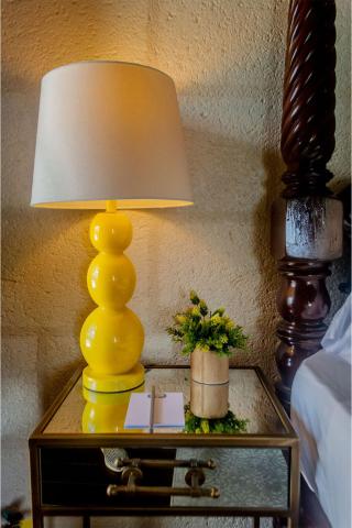 The image shows a brightly colored yellow table lamp with a curvy base and a white lampshade, positioned on a wooden nightstand