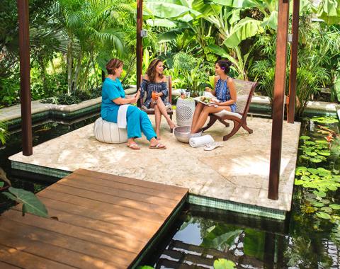 The image shows three women sitting and chatting in a serene outdoor setting that includes a small pond with lily pads