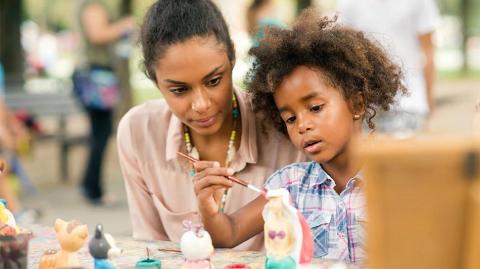 The image shows a young woman and a little girl engaging in a craft activity outdoors