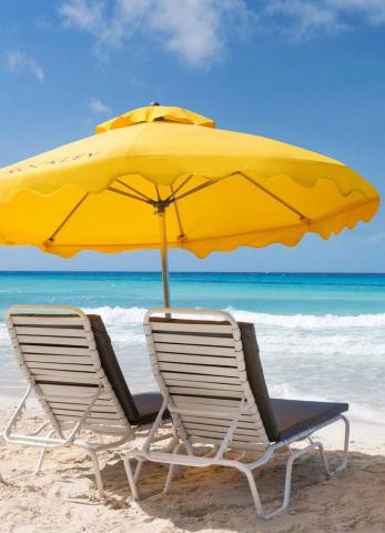 The image displays a beautiful beach scene featuring a large yellow umbrella and two white and gray lounge chairs