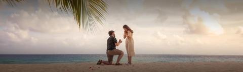 This image captures a romantic moment on a beach where a man is kneeling on one knee, likely proposing to a woman