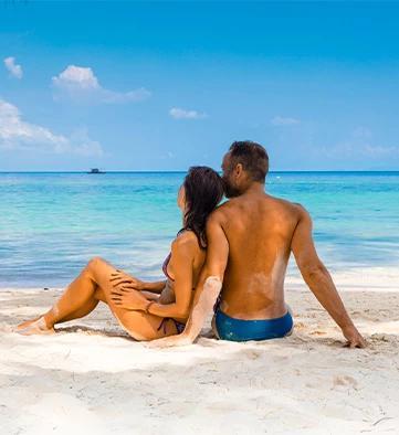 The image shows a couple sitting on a sandy beach, enjoying a sunny day by the clear blue sea