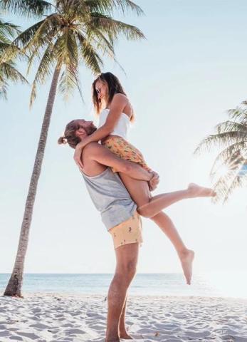 This image features a joyful scene at a beach with a man lifting a woman in his arms