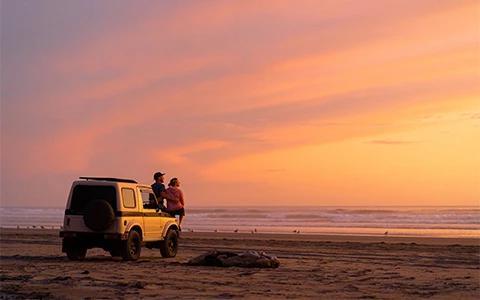 This image shows two people sitting on the tailgate of an SUV, parked on a sandy beach at sunset