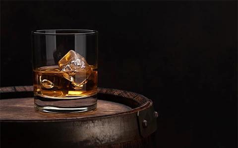 The image shows a glass of whiskey with ice cubes, resting on a wooden barrel