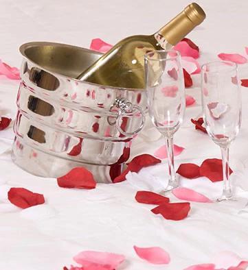 The image features a romantic setting that includes a silver ice bucket with a bottle of wine, two empty glass flutes, and scattered red rose petals, all placed on a white textured background