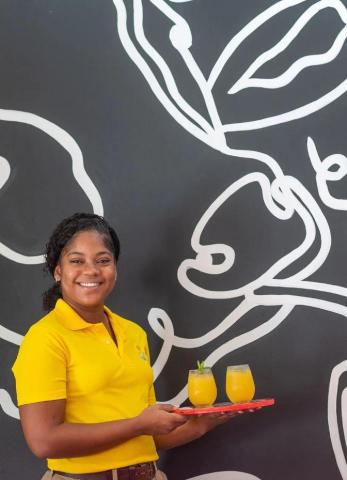 This image shows a cheerful woman wearing a yellow polo shirt, standing in front of a dark grey wall with a white line-art drawing