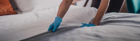 The image shows a person making a bed and wearing blue gloves, probably indicating that they are performing some sort of cleaning or housekeeping work