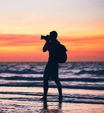 This image features a silhouette of a person standing in shallow ocean water at sunset, taking a photograph
